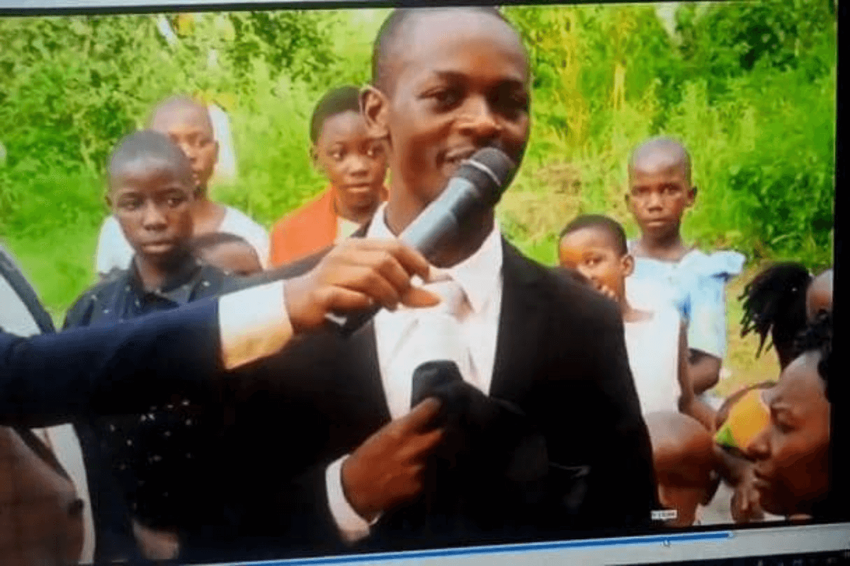Ambrose Katende speaking into the microphone. His sponsor student is standing behind hiim in the black shirt. 